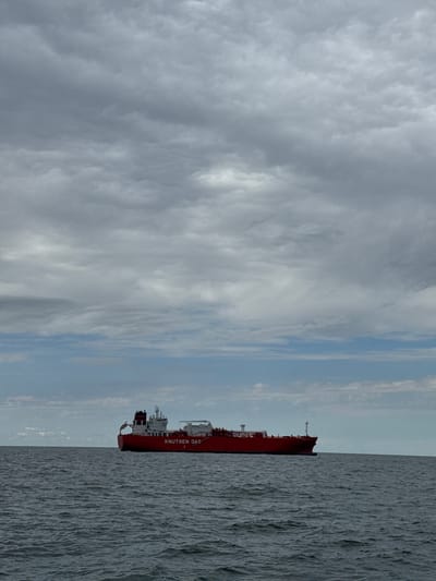 The image captures a large red tanker ship at sea under a vast, partly cloudy sky. The vessel is oriented horizontally, with its bow extending to the left and its stern, featuring a prominent superstructure, towards the right. The ship's hull is a deep red, contrasted by white superstructures. The name "KNUTSEN OAS" is clearly legible in white capital letters along its side, followed by what appears to be the numeral '1'.

The sea, occupying the lower half and right side of the frame, is a dark, somewhat turbulent expanse of water, showing numerous ripples and small waves. The sky above, filling the upper half and left side, is dominated by varying shades of gray clouds, ranging from dense, textured formations to lighter, more translucent areas. Patches of clear blue sky are visible through breaks in the clouds, particularly towards the left horizon.

The scene depicts a daytime setting, with a generally overcast but not dark sky, suggesting diffused light. There is no specific activity or people visible. The overall impression is one of a large maritime vessel in the open waters. The location context for this scene is Marina di Ravenna, Italy.