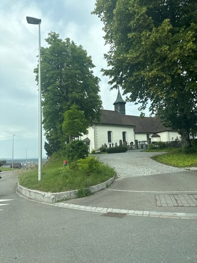 The image shows a white church with a steeple, situated in the city of Kreuzlingen, Switzerland. The church has a dark-tiled roof and a graveyard in front of it. The church is surrounded by trees and greenery. A cobblestone pathway leads to the church entrance. In the foreground, there is a road with a speed limit sign and a street lamp. The sky is overcast. The overall scene depicts a quiet, rural setting with a focus on the church and its surroundings.