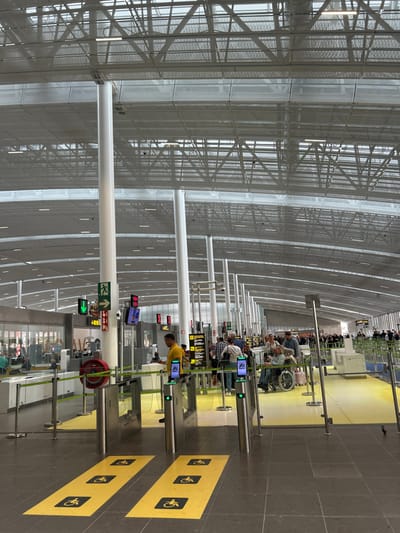 The image shows a section of the Gran Canaria Airport, located at latitude: 28.047, longitude: -16.58, in the city of Granadilla, Spain. It captures the airport's modern architecture with a high, white, and grey ceiling featuring a mesh of metal beams and a large open space. The scene depicts a line of people waiting to pass through security at a large gate. People are visible walking towards the gate, holding their luggage, or sitting in a wheelchair while being pushed by another person. Security personnel are also visible behind the gate, which is marked with a yellow line and a yellow and red rope. Two metal security gates are visible, along with two digital screens. Yellow stickers with the symbol for handicapped people are placed on the floor, indicating accessible paths.