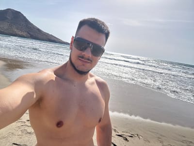 A man with short dark hair, a beard, and sunglasses stands on a beach in Níjar, Spain, taking a selfie. He is shirtless and wearing dark sunglasses. The beach has light-colored sand, and the ocean has moderate waves with whitecaps. A large, barren, dark-colored mountain is visible in the background on the left side of the image, with the sky above being clear and light blue. The sun is shining, casting shadows and making the water sparkle.