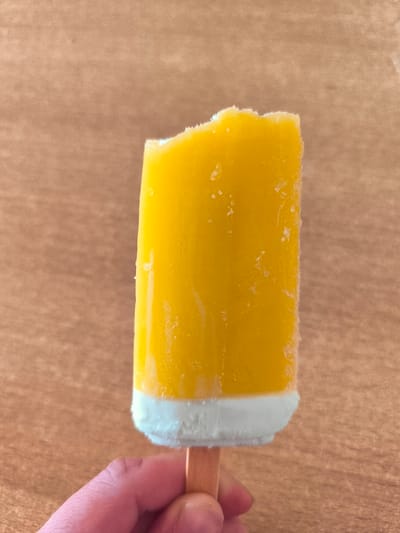 A close-up, eye-level shot captures a person's hand holding a half-eaten, yellow popsicle with a white bottom. The popsicle has a bite taken out of its top, revealing a slightly rough texture. The background is a textured, light brown surface, resembling wood. The scene is likely indoors, given the lighting. The exact location in Wervershoof, Netherlands cannot be determined from the visual information provided.
