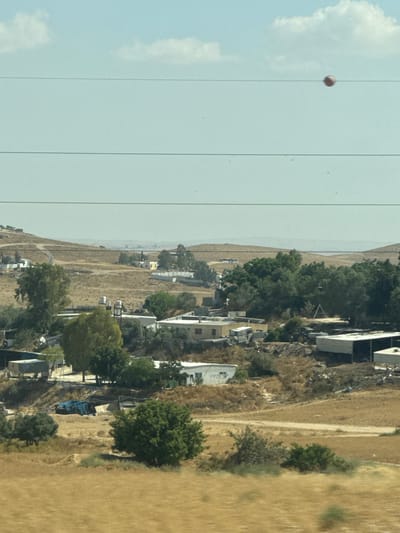 The image presents a view of Al-Qasoum Regional Council, Israel, characterized by arid, dry terrain transitioning into sparse greenery and a scattering of structures resembling houses or dwellings. Power lines cut across a pale blue sky containing clouds. The overall landscape suggests a rural or semi-rural environment. A single, round object is suspended on a power line.