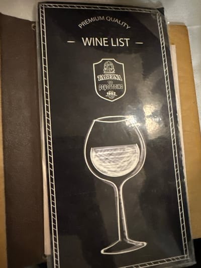 The image displays a close-up, slightly angled shot of a black wine list cover, indicative of a restaurant or tavern setting. The cover features white or silver graphics and text against its dark background, framed by a decorative double-line border pattern along the top, bottom, and left edges.

The primary subject is the wine list cover itself. On the right side, a stylized outline of a wine glass is depicted, with its bowl partially filled with a textured, possibly frosted, white pattern. On the left, rotated 90 degrees counter-clockwise, the words "WINE LIST" are prominently displayed, with "PREMIUM QUALITY" written above it, also rotated.

In the center-left of the cover is a shield-shaped emblem or logo. Within this emblem, the text "TAVERNA", "TO", and "ΡΟΥΤΟΥΚΟ" (Routouko) are visible, along with the year "1982". This identifies the establishment as "Taverna to Routouko" and indicates its founding year, providing a strong location cue for a Greek dining place, specifically in Agios Dimitrios.

The wine list appears to be resting on a dark brown surface, possibly another menu or a table, with a lighter brown object visible underneath it. The lighting suggests an indoor environment. No people are visible, and there is no discernible activity beyond the presentation of the menu. The overall impression is one of a formal yet inviting dining experience.