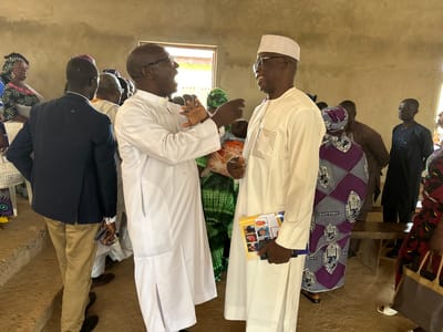 The image depicts an indoor gathering of people in what appears to be a community hall or church in Kufana, Nigeria. Two men in the foreground are engaged in conversation. The man on the left wears a white religious garment and glasses, gesturing with his hands. The man on the right, also in white attire and a white cap, smiles and holds a booklet or pamphlet. Other individuals are visible in the background, some wearing traditional African clothing and head coverings. The setting is rustic, with bare walls and simple wooden benches. The lighting suggests it is daytime. The overall mood seems to be social and perhaps celebratory, given the smiles and interaction. Visible text on the booklet held by the man on the right includes what appears to be "BAPTIST" and possibly "WOMEN". The walls have a rough, textured finish, and there is a window with natural light streaming through. The floor is a dirt or concrete surface. The event appears to be a community or religious gathering.