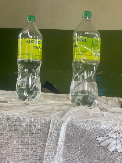Two clear plastic water bottles with green caps stand upright on a beige, textured tablecloth. Both bottles contain clear liquid and have yellow labels with text. The bottle on the left is slightly closer to the viewer. The background is a dark green wall.
