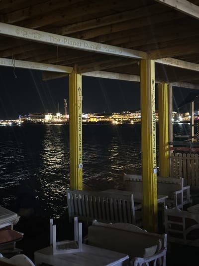 The image depicts an outdoor dining area, likely at night, in Dahab, Egypt. The restaurant or cafe is situated on or near the water, as suggested by the illuminated reflections on the water's surface. Structural wooden beams, labeled "The Planet Dahab" in English, support an overhead structure or roof. White chairs and tables are arranged around the space. The image is taken at an oblique angle and rotated counterclockwise by 90 degrees. The city lights of Dahab are visible in the distance across the water.