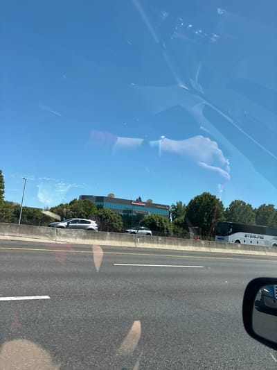 The image captures a bright, sunny day in Rancho Cordova, California, as seen from the perspective of a vehicle traveling on Highway 50 East. The foreground is dominated by the asphalt of the highway, marked with white lane lines and a yellow center line. In the mid-ground, a concrete barrier separates the highway from an elevated area. Beyond the barrier, a silver SUV is parked near a building with the "Bank of America" logo visible. Further to the right, a white Sterling tour bus is driving on the highway. The background consists of lush green trees and a clear blue sky. Reflections on the vehicle's windshield obscure parts of the scene, including what appears to be a person's hand.