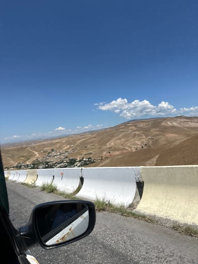 An over-the-shoulder, eye-level shot captures a rural road in Беш-Мойнок, Kyrgyzstan, on a clear, sunny day. The perspective is from inside a vehicle, looking out past a side-view mirror. The road, paved with dark asphalt, curves gently to the right. A concrete barrier, painted white and yellow, separates the road from a steep, arid hillside. The hillside is dotted with a village of scattered white houses and is crisscrossed by unpaved roads. In the distance, rolling, dry, brown hills stretch towards the horizon under a vast blue sky with scattered white clouds. The side-view mirror on the left reflects the interior of the car and a person wearing a blue patterned shirt. The mirror itself shows signs of rust and peeling paint.