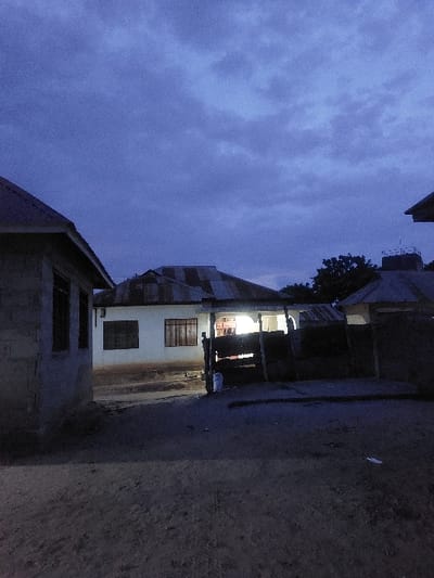 The image depicts an exterior scene in Mwandege, Tanzania, at dusk or early night. The sky overhead is dark blue to purple, with visible cloud formations. The ground in the foreground and mid-ground consists of unpaved dirt. On the left, a partial view of a structure with unpainted concrete block walls and a dark sloped roof is present, showing dark window openings. Centrally positioned is a single-story, light-colored building with a corrugated metal roof. This central building features two windows with dark frames and horizontal grates. An interior artificial light source brightly illuminates a section of its facade, likely a porch or open doorway, casting a yellow glow. A dark wooden fence structure or gate stands directly in front of this illuminated area, with a white bucket visible near its base on the left. To the far right, a partial view of another building with a corrugated metal roof is discernible, with dark tree foliage behind it. No human figures, specific actions, or interactions are visible within the frame.