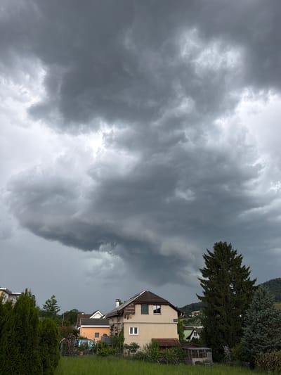 In Gelterkinden, Switzerland, a dramatic sky filled with dark, heavy storm clouds dominates the scene. The clouds appear dense and turbulent, suggesting an impending downpour. Below the ominous sky, several houses are visible. The largest house in the foreground is a light beige, multi-story building with a dark brown roof. It has a chimney and visible windows, one of which is a small square window on the upper floor. To the left of this house is a smaller, brightly colored orange house. Further left, a section of another building with a white facade is visible. In the mid-ground, a light blue vehicle is parked behind a fence adorned with pink flowers. A large, dark green evergreen tree stands prominently to the right of the main house, with another similar tree to its right. In the background, rolling hills with scattered houses and trees suggest a suburban or semi-rural setting. The foreground consists of a green grassy field.