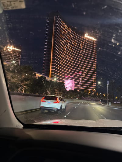 The image shows a nighttime view of the Encore hotel in Las Vegas, Nevada, captured from inside a vehicle. The hotel's illuminated façade, with its rows of lights and the "Encore" sign, dominates the left side of the frame. A street runs alongside the hotel, with another vehicle visible. The scene is viewed through a car window. There are no people visible.