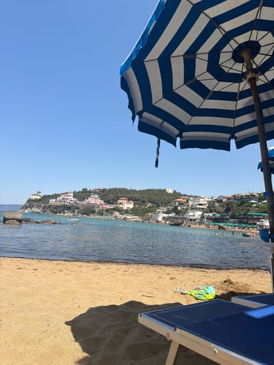 The image shows a view of the beach and sea in Castiglioncello, Italy. In the foreground, there is a sandy beach with two blue beach chairs and a blue and white striped beach umbrella. A green plastic bag lies on the sand between the chairs. The sea is visible in the middle ground, with several people swimming and floating in the water. In the background, there is a town with buildings on a hill overlooking the sea. The sky is clear and blue. The overall scene depicts a sunny day at the beach.