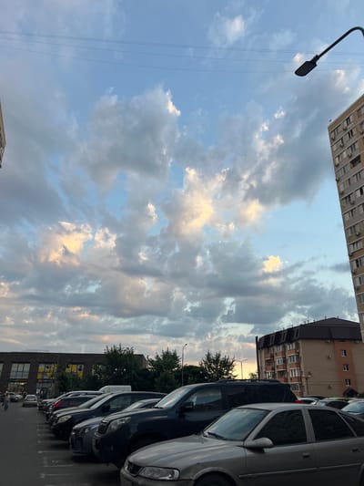 The image captures a parking lot in Belozernyi, Russia, with a backdrop of an overcast sky featuring scattered clouds tinged with sunset colors. In the foreground, several cars are parked in neat rows. To the right, a tall apartment building with numerous windows and air conditioning units is visible. To the left, a lower building with a dark facade, large windows, and a sign that appears to say "CI.LAB", suggesting a commercial establishment like a laboratory or shop, is present. Trees are interspersed between the buildings and the parking lot. A streetlamp arcs over the parking area. The overall scene depicts an urban residential and commercial area during dusk.