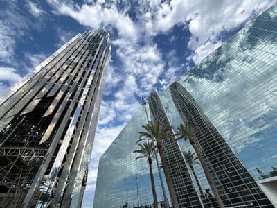 The image shows the exterior of a modern building complex in Garden Grove, California, United States of America. In the foreground, a tall, angular building with a glass facade reflects the sky and clouds. To the left, another structure, appearing to be under construction or renovation, features a metallic, ribbed exterior. Several palm trees are visible in front of the glass building, their reflections also captured in the facade. The sky is blue with scattered white clouds, indicating fair weather. The perspective is from a low angle, looking upwards at the buildings. There are no people visible in the scene.