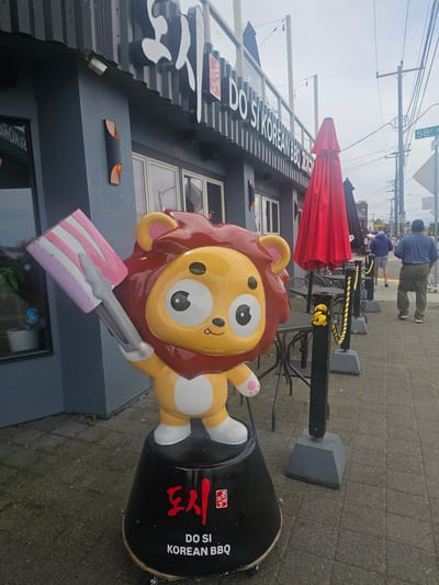An exterior shot shows the front of a business named "DO SI KOREAN BBQ" located in Seattle, USA. The building is dark gray with large windows. A cartoonish lion statue, yellow with a red mane, stands outside, holding a pink and white striped popsicle. The lion stands on a black base with Korean characters and "DO SI KOREAN BBQ" written on it. Red and black umbrellas are set up near tables outside the restaurant. Two men are walking away from the camera on the sidewalk. A street sign for "58th Ave" is visible. The sky is overcast.