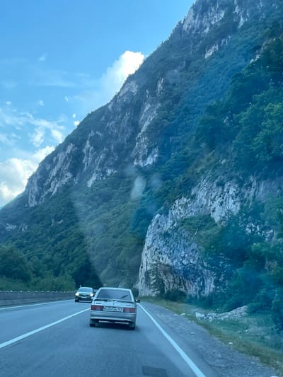 The image captures a scenic drive along a mountainous road, with lush green vegetation covering the slopes of a towering rock face. The sky above is a clear blue with a few scattered clouds. In the foreground, a silver car is driving on the asphalt road, with another car visible further ahead. The road appears to be a multi-lane highway, with white lines marking the lanes. The overall impression is one of natural beauty and a journey through a picturesque landscape. The location in the city of Balta, Russia is not explicitly mentioned in the image.