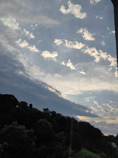 This is a vertical, eye-level, full-shot photograph of a forested hillside under a cloudy sky. The sky occupies the top two-thirds of the image and transitions from a light blue at the top to darker grays and whites nearer the horizon. Small, scattered white clouds are visible, interspersed with larger, darker gray clouds that form a diagonal band across the upper left portion of the sky. The lower third of the image depicts a densely forested hillside. The trees are dark green and silhouetted against the sky, with some lighter green foliage visible at the bottom. A small, reddish-brown tiled roof of a building is partially visible peeking out from the trees on the right side of the hillside. The overall lighting suggests it is either early morning or late afternoon. The scene is located in Bolshoy Sochi, Russia, likely a residential area with a natural landscape.