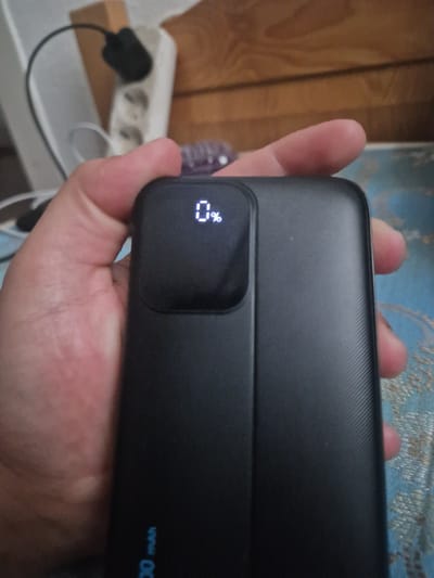 A hand is holding a black power bank. The power bank has a small digital display that shows "0%". The background is blurred and shows a wooden surface, an electrical outlet, and a wire. The power bank has text in blue that reads "00 mAh". The image does not provide information about the city of Überlingen, Germany.