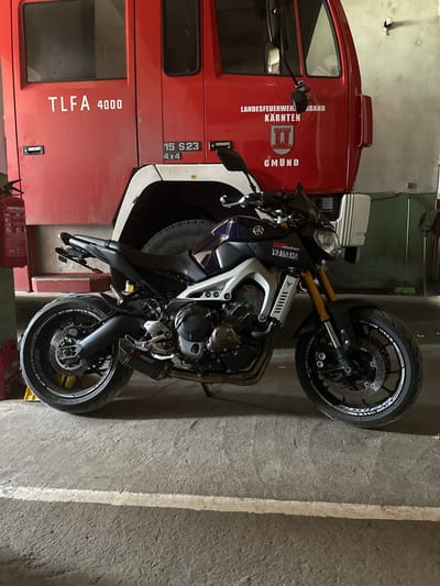 A red fire truck is parked behind a dark purple and silver Yamaha motorcycle in what appears to be a garage or workshop. The fire truck has text on its side including "TLFA 4000", "15 S23 4X4", "LANDESFEUERWEHRBAND KÄRNTEN", and "GMÜND". The motorcycle has "YAMAHA" and "MT-09" logos visible. A fire extinguisher is mounted on a stand to the left of the motorcycle, and a broom is leaning against the wall to the right. The floor is concrete, with a white line painted across it. The overall lighting suggests an indoor setting. The image does not depict the city of Valchedram, Bulgaria.