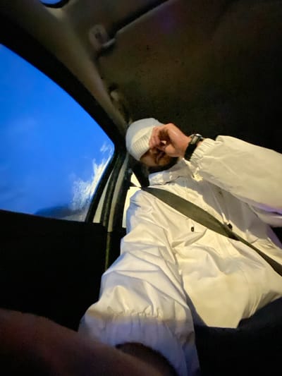 An upside-down view inside a car captures a person wearing a white jacket and cap, secured by a seatbelt. The sky is visible through the open sunroof. The person's wrist reveals a black watch. Dark upholstery and interior elements surround the subject, creating a confined setting. The image's orientation offers a unique perspective, emphasizing the person's position within the vehicle and the outside world.