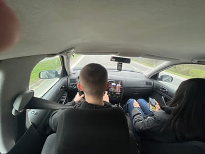 The image depicts the interior of a vehicle, viewed from a rear passenger seat, in motion on a road in Zagore, Bulgaria. In the driver's seat, a male with short hair is operating the vehicle, with both hands on the steering wheel. He is wearing a dark top, and a seatbelt is visible across his left shoulder. In the front passenger seat, a female with long dark hair is seated, wearing a dark jacket and blue jeans. She is holding a smartphone in both hands, actively engaged with its screen, and her feet are elevated, resting against the dashboard. A seatbelt is not visibly secured on her. The car's dashboard is dark, featuring air vents, climate controls, and a rearview mirror with a black air freshener hanging from it. Several colorful cans or bottles are positioned on the central console below the dashboard. Through the windshield and side windows, a two-lane road with a white broken center line and a solid white shoulder line is visible, flanked by green grass and vegetation under an overcast sky. A finger belonging to the person taking the photograph is visible in the upper left corner.