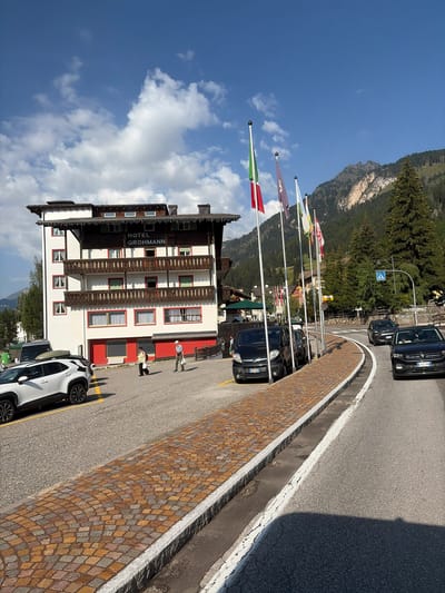 In Campitello di Fassa, Italy, a white building with the sign "HOTEL GROHMANN" is visible. The hotel has several floors with balconies and red-trimmed windows. In front of the hotel, there are two pedestrians, one wearing a yellow shirt and the other a white shirt and hat, walking on a paved area. Several cars are parked along the side of the road, including a white SUV, black vans, and black cars. Flags are displayed on flagpoles in front of the hotel. The road curves to the right, with a pedestrian crossing sign visible in the distance. Mountains covered with trees form the backdrop of the scene, with a clear blue sky and scattered clouds above.