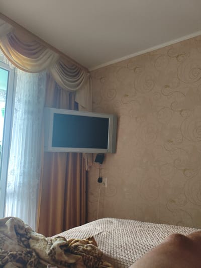 This image shows a bedroom scene. A television is mounted on a wall covered with a patterned wallpaper. There are curtains on the left side of the image, and a bed with blankets is in the foreground. The overall impression is that of a functional, lived-in space.