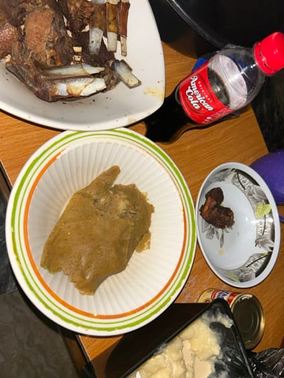 In the city of Debasco, Nigeria, a wooden table displays a prepared meal. A large white oval platter at the upper left holds multiple pieces of dark brown, roasted rib meat. Below this, a white bowl with green and orange stripes contains a substantial portion of light brown, mashed food. To the right, a small white bowl with a floral pattern holds a single piece of dark brown roasted meat. A partially compressed clear plastic bottle of "Original American Cola" with a red cap is positioned near the roasted ribs. In the lower right, a black container partially reveals a creamy, off-white, lumpy substance, with a metal can visible next to it. No individuals are present in the scene.