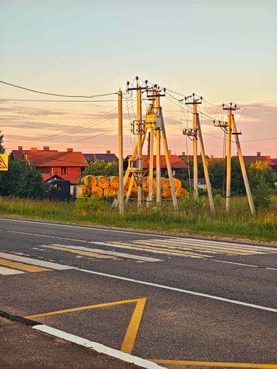 This image depicts a rural road scene in the town of Kurovo, Russia. The foreground features a crosswalk with yellow and white markings on an asphalt road. To the right, a curb with black and white paint borders the road. In the middle ground, a grassy verge separates the road from a collection of large, round hay bales stacked on a flatbed truck. Behind the hay bales, several power line poles with electrical equipment are visible. Further in the background, a cluster of red-roofed houses is nestled among green trees. The sky above is a pale blue with wispy, pink-tinged clouds, suggesting either dawn or dusk. A yellow warning sign with a black silhouette of a pedestrian is visible on the left side of the road. The overall impression is one of a quiet, rural setting during a calm time of day.