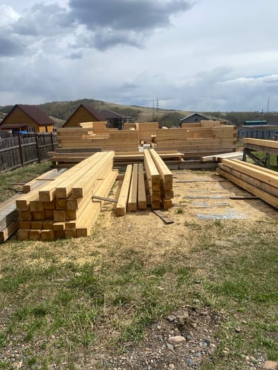 The image depicts a construction site or lumberyard located in Solontsy, Russia. Stacks of freshly cut wooden beams and planks are the primary subjects, arranged on a grassy area with some scattered stones. The sky is overcast with heavy clouds, suggesting possible inclement weather. In the background, there are residential houses and a tree-covered hill, characteristic of the Solontsy landscape. The scene is devoid of human presence, focusing on the material prepared for building.