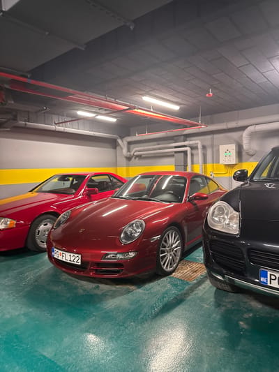 The image shows a section of an indoor parking garage. There are three cars visible. The closest car to the camera is a red Porsche, with a license plate that reads "PG FL122". Behind the Porsche is another red car. Next to the Porsche is a black SUV-type vehicle with a license plate beginning "MNE". The parking garage floor is painted in green, with yellow stripes on the walls indicating parking spots. Pipes and utility infrastructure are visible on the ceiling and walls of the garage. The location is not explicitly identified as Katun Bačko brdo, Montenegro, but the license plate "PG" is a likely indicator that the cars are registered in Montenegro. The image lacks any visible people, subjects, actions or interactions.