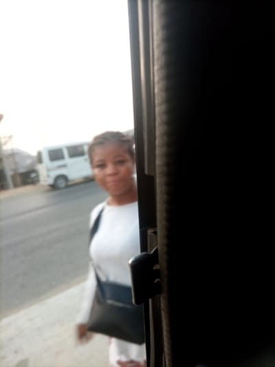 The image displays an outdoor scene in the city of Obehie, Nigeria, viewed from an interior perspective through an opening. A dark vertical structure, featuring a black latch mechanism, frames the right side of the image. A female individual is positioned in the left portion of the frame, out of focus, wearing a white long-sleeved top and a black shoulder bag across her body. Her hair is braided, and her face is oriented towards the camera. In the blurred background, a white minivan is visible on a paved road. Distant buildings and structures are indistinct, beneath a bright, uniformly lit sky.