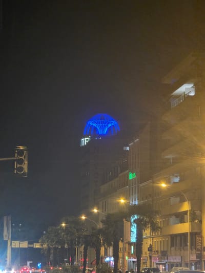 This is a night-time, low-angle, vertical photograph of an urban street scene in Montrouge, France, dominated by the illuminated dome of a building at the center, glowing bright blue. A traffic light is visible on the left side of the frame. Adjacent buildings line the street, with multiple floors and balconies, many windows are illuminated, casting a warm, yellow glow. The building with the blue dome has white lettering that appears to read "ITP". Below this, on another building, a green neon sign reads "ibis styles". A tall street lamp with a bright yellow light is prominent on the right, its light rays fanning out across the image. Several palm trees are sparsely planted along the sidewalk, with their silhouettes visible against the illuminated buildings. Traffic is visible in the foreground, with red taillights suggesting a slow or stopped flow of vehicles. The overall atmosphere is that of a city street at night, with artificial lighting creating a somewhat hazy or foggy effect. An "AVIS" sign is visible on a building towards the right.