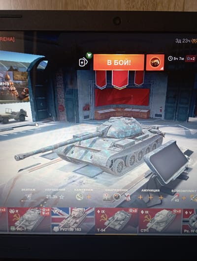The image depicts a screenshot of a video game, likely a tank combat simulator, displayed on a laptop. The game interface shows a virtual garage with a detailed 3D model of a tank as the main subject. Text labels in Russian are visible, indicating game options such as "ЭКИПАЖ" (Crew), "УЛУЧШЕНИЯ" (Upgrades), "КАМУФЛЯЖ" (Camouflage), "СНАРЯЖЕНИЕ" (Equipment), "АМУНИЦИЯ" (Ammunition), and "БОЕКОМПЛЕКТ" (Shells). Below the main tank, a selection of smaller tank icons is displayed, each representing a different in-game vehicle. The laptop itself is placed on a wooden surface, suggesting it is being used in a domestic or casual setting. There is no discernible connection to the city of Esik, Kazakhstan, or any specific real-world location. The scene is entirely digital and contained within the game's virtual environment.