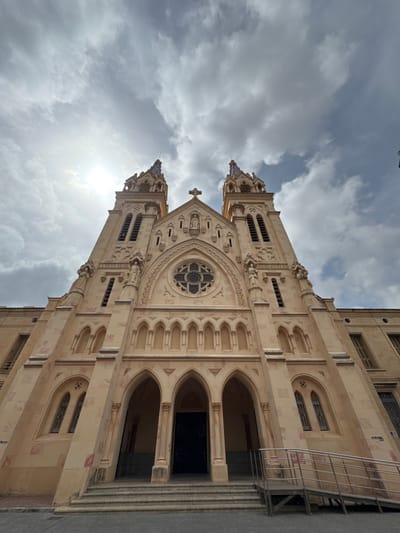 This is a vertical, low-angle, outdoor photograph of a large, pale stone building, possibly a church or cathedral, under a cloudy sky. The building features Gothic architectural elements, including pointed arches, ornate stone carvings, and a rose window above the entrance. The facade is divided into multiple stories with repetitive window patterns and decorative stone trim. A tall, slender tower with a cross on top is visible on the left side of the building, and another similar tower is partially visible on the right. Sunlight breaks through the clouds in the upper left portion of the image, casting a bright glow. The building appears to be made of light-colored stone with some reddish-brown staining or inclusions. The scene is devoid of people, creating a sense of stillness and grandeur. There is a metal staircase attached to the right side of the building, leading up to an upper level. The sky is filled with heavy, grey clouds, suggesting overcast weather. The orientation of the image is such that the building appears to be leaning to the left.