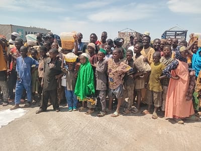 The image shows a group of 35 people standing on a paved road in Katabu, Nigeria, located at coordinates 10.71 latitude and 7.514 longitude. They are primarily men and boys, with a few women and girls present. Most of the people are wearing casual clothing, including t-shirts, jeans, and traditional robes. Some individuals are holding buckets, possibly containing food or water. Several trucks are parked in the background, and there is a container labeled "MAERSK" visible. The scene appears to be an informal gathering, with people interacting and talking amongst themselves. The sky is blue with some white clouds, indicating a sunny day. The ground is paved and has some white painted lines. The general atmosphere is one of casual interaction and possibly a sense of anticipation or waiting.