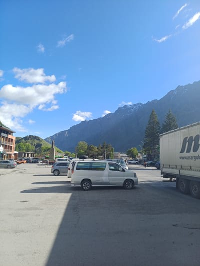 The image depicts a sunlit scene in a town square, likely in Stepantsminda, Georgia, with a prominent backdrop of rugged, snow-capped mountains. The sky is a clear blue with scattered white clouds. In the foreground, a white minivan is parked on a paved surface. To the right, a large white truck with the logo "m www.marguli" is visible, partially obstructing the view. The town square is populated with several other vehicles, including cars and a grey SUV. A building with orange and brown accents stands on the left, and a statue of a person is centrally located in the mid-ground. Green trees dot the landscape, and more buildings are visible in the distance. The overall impression is that of a bustling, somewhat rural, mountain town on a bright day.