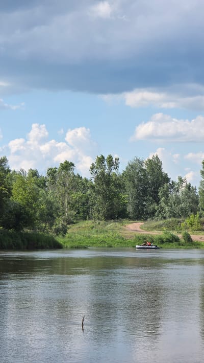 The image shows a wide river with a calm surface reflecting the sky and trees. On the opposite bank, there is a dense line of lush green trees. A dirt path winds through the grass on the bank, leading towards the trees. In the middle of the river, a small inflatable boat with two people in orange life vests is visible. The sky above is partly cloudy, with large grey clouds interspersed with patches of blue sky and smaller white cumulus clouds. The scene appears to be set in a natural, rural environment.