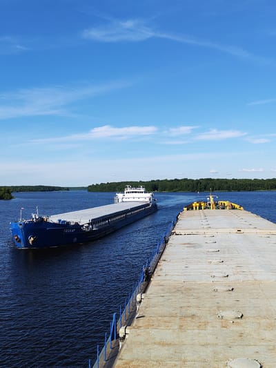 This media file captures a bright, sunny day on a wide river, likely in Nova, Russia. The scene is dominated by two large barges moving on the deep blue water under a clear sky dotted with wispy white clouds. The sun is high, suggesting it is midday.
In the foreground, a flat-topped barge, mostly gray and weathered, stretches out of view to the right. Its surface is marked with several circular hatches and a blue railing runs along its edge. In the middle ground, a longer blue barge, the "СЕСКАР" (SESKAR), is moving towards the viewer. It has a white superstructure at its stern, and a Russian flag flies from its bow. This barge appears to be carrying cargo under its large, flat deck. In the distance, a dense line of green trees forms the shoreline on both sides of the river, indicating a rural or forested area. The water is calm with gentle ripples. There are no people visible in the image. The overall mood is serene and evokes a sense of calm industrial activity.