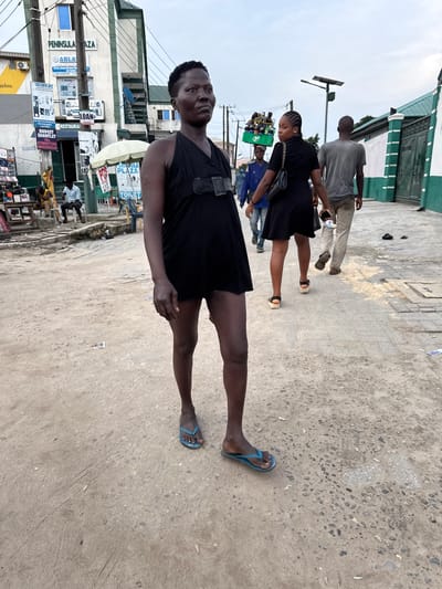 The image depicts a street scene in Lagos, Nigeria, likely in the Ajegunle area, characterized by a mix of commercial buildings and informal market activities. In the foreground, a woman with dark skin and short hair stands facing slightly to the right, wearing a black sleeveless dress and blue flip-flops. Her hands are at her sides, and she appears to be looking off into the distance. Behind her, a woman in a black dress and platform sandals walks away from the camera, carrying a black handbag. Further in the background, a man in a grey t-shirt and light-colored pants also walks away. Other individuals and a vendor with goods on their head are visible in the mid-ground, along with signs for "PENINSULA PLAZA" and "PUBLIC TOILETS." The ground is paved with concrete and dirt, with some debris scattered around. The overall atmosphere suggests a busy urban environment with a pedestrian-focused streetscape.