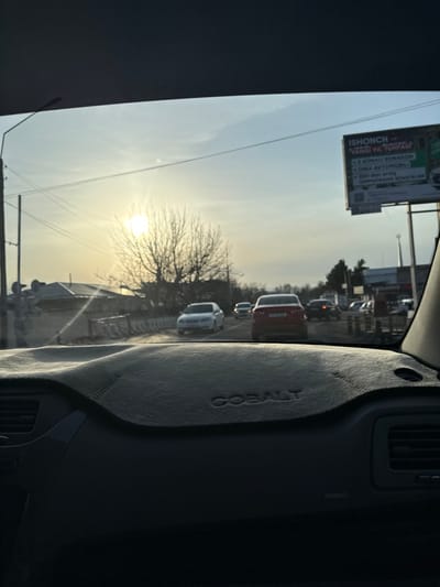 Captured from inside a car, the view shows a busy road with multiple vehicles. The sun is low, shining through a bare tree, creating a hazy, bright atmosphere. A sign with text in a foreign language stands prominently on the roadside. Part of the car's interior, including a dashboard cover with the word "COBALT," is visible, adding context to the scene.