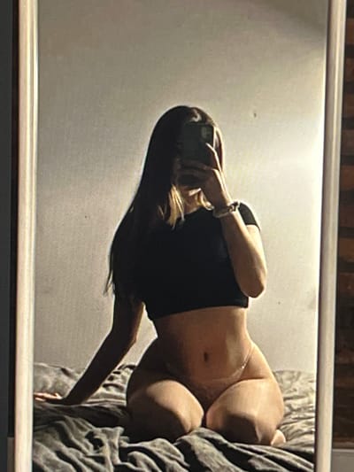 The image shows a person taking a mirror selfie in what appears to be a bedroom. The person is wearing a black crop top and light-colored underwear. They are sitting on a bed with grey bedding. The background is a plain wall, and the mirror frame is visible on the right side of the image. The lighting suggests an indoor setting, possibly at night or in a dimly lit room. There is no information in the image to suggest it is located in Pruszcz Gdański, Poland.