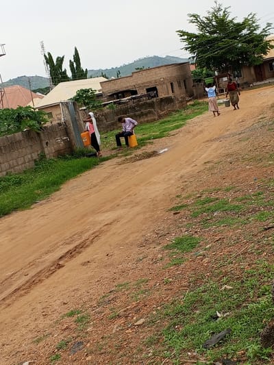 In Ashata, Nigeria, an outdoor scene depicts several individuals engaged in various activities. A person wearing a white hijab and a pink top stands near a water source, holding an orange jerrycan. To their right, a young man in a light purple shirt and dark pants sits on a yellow jerrycan. Further in the background, two women are walking, one carrying a green bowl on her head and the other holding a bucket. They are on a dirt road that winds through a residential area with brick buildings and a large tree. The environment appears dry, with scattered green vegetation and a cloudy sky.