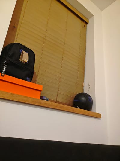 A low-angle, interior shot shows a window ledge with various objects resting upon it. The window behind is covered with horizontal, tan-colored blinds. On the ledge, from left to right, are an orange rectangular box, a black backpack with a Nike tag and a "JUST DO IT." keychain, a small pack of cigarettes or mints, and a black soccer ball with blue accents. The wall to the right of the window is white. The bottom portion of the image shows a black, upholstered surface. The location is likely a private residence in an unknown location within Swindon, United Kingdom.