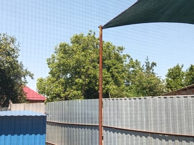 This media file captures an outdoor scene on a bright, sunny day, likely in Tbilisskaya, Russia. The primary subjects are a tall, brown metal pole supporting a dark green shade sail that stretches across the top of the frame. The sail is secured to the pole with yellow ties.

In the background, a dense canopy of lush green trees fills the mid-ground, indicating a warm season. Behind the trees, a building with a red roof is partially visible, suggesting a residential or perhaps a rural setting. A corrugated metal fence runs horizontally across the lower half of the image, dividing the foreground from the background. In the lower left, a portion of a blue metal structure, possibly a shed or a storage unit, is visible.

The overall impression is one of a clear, bright day. The sunlight casts shadows, especially on the corrugated fence, further emphasizing the time of day as being midday or early afternoon. There is no visible text in the image. The scene suggests a peaceful, possibly a private yard or a communal area being shaded from the sun.