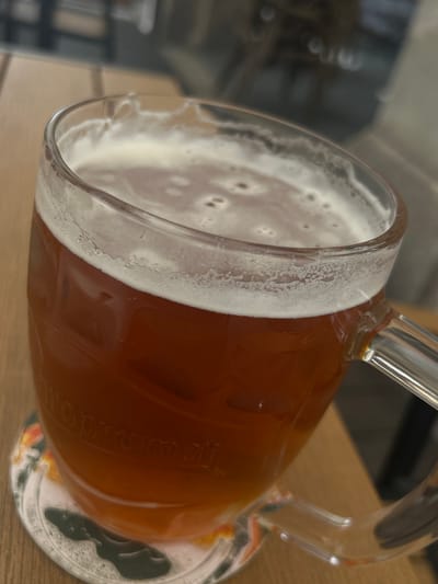 A close-up, high-angle shot captures a glass beer mug filled with a reddish-brown beverage, likely a lager or ale, resting on a light-colored wooden table. A thick layer of white foam tops the drink, and "lacing" from the foam adheres to the inner wall of the clear glass mug. The mug features a prominent handle on the right side. Beneath the mug is a circular coaster with an indistinct, partially obscured pattern, including a dark, irregular shape. Faintly embossed or printed on the side of the glass, the word "Prazdroj" is visible, rotated, suggesting a connection to Pilsner Urquell, a prominent Czech beer brand. The background is softly blurred, showing only vague shapes, possibly shelves or other indoor elements. The image is taken indoors, with no specific time of day or weather discernible. The overall scene suggests a casual setting, possibly a pub, restaurant, or home, consistent with the given context of Liberec, Czechia.