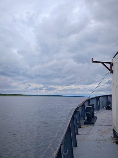The image shows a view from the deck of a ship on a river in Zotino, Russia. The sky is overcast with gray clouds. The river is wide and calm, with a tree-lined shore in the distance. On the deck of the ship, there are bollards and railings. A white cargo container is visible on the right side of the image. The scene is likely captured during daytime, given the ambient light. There are no people or animals visible in the image.