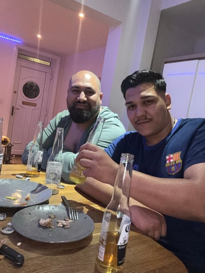 Two men are seated at a wooden table, with several bottles of Corona beer and dirty plates of food scattered around them. The man on the left has a shaved head and a beard, wearing a light blue hoodie. The man on the right has dark hair and a mustache, wearing a blue t-shirt with the FC Barcelona crest. The background shows a doorway and a wall with subtle lighting. The setting appears to be a casual indoor gathering.