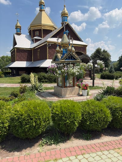 A bright, sunny day in Triitsia, Ukraine, features a traditional Ukrainian Orthodox church with golden domes and dark brown shingled roofs. The church is adorned with blue and gold accents, including crosses on its spires. In the foreground, a paved walkway with red and beige diamond-shaped tiles borders a meticulously maintained garden. Lush green, rounded bushes and taller, wispy green plants are arranged along the path. Hanging flower baskets overflow with white and purple blossoms, and a tall plant with a spikey stalk and white flowers stands out in the garden. The scene is peaceful and serene, capturing the beauty of a religious site and its surrounding landscape on a clear day.
