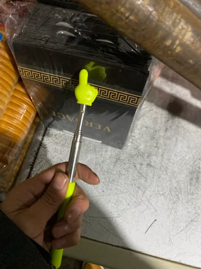 A hand holds an extendable pointer with a yellow plastic thumbs-up attached to the end. The pointer rests on top of a stack of black Versace-branded napkins in plastic packaging. Beneath the napkins are stacks of round, golden-brown cracker-like snacks, also in plastic wrap. A cylindrical metal object is partially visible to the left. The background consists of a scratched, white, flat surface.